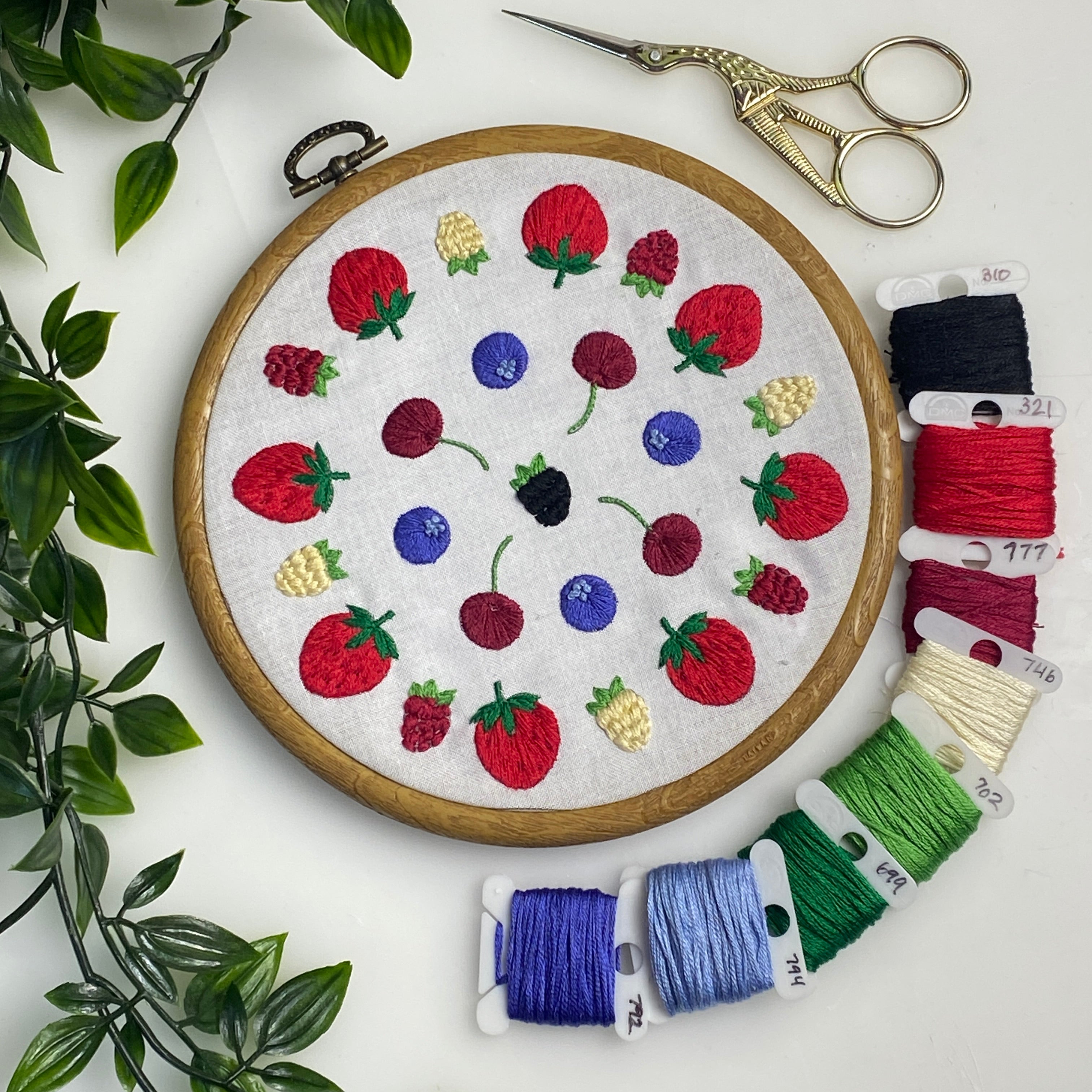 A round embroidery hoop displaying a colorful embroidered fruit pattern on white fabric, arranged on a white surface with decorative green leaves, gold scissors, and multiple spools of thread in red, blue, green, purple, and cream.