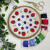 A round embroidery hoop displaying a colorful embroidered fruit pattern on white fabric, arranged on a white surface with decorative green leaves, gold scissors, and multiple spools of thread in red, blue, green, purple, and cream.