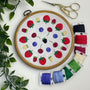 A round embroidery hoop displaying a colorful embroidered fruit pattern on white fabric, arranged on a white surface with decorative green leaves, gold scissors, and multiple spools of thread in red, blue, green, purple, and cream.