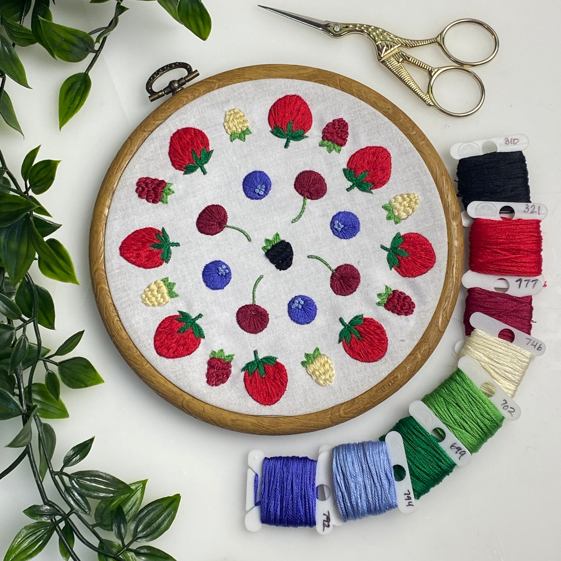 A round embroidery hoop displaying a colorful embroidered fruit pattern on white fabric, arranged on a white surface with decorative green leaves, gold scissors, and multiple spools of thread in red, blue, green, purple, and cream.
