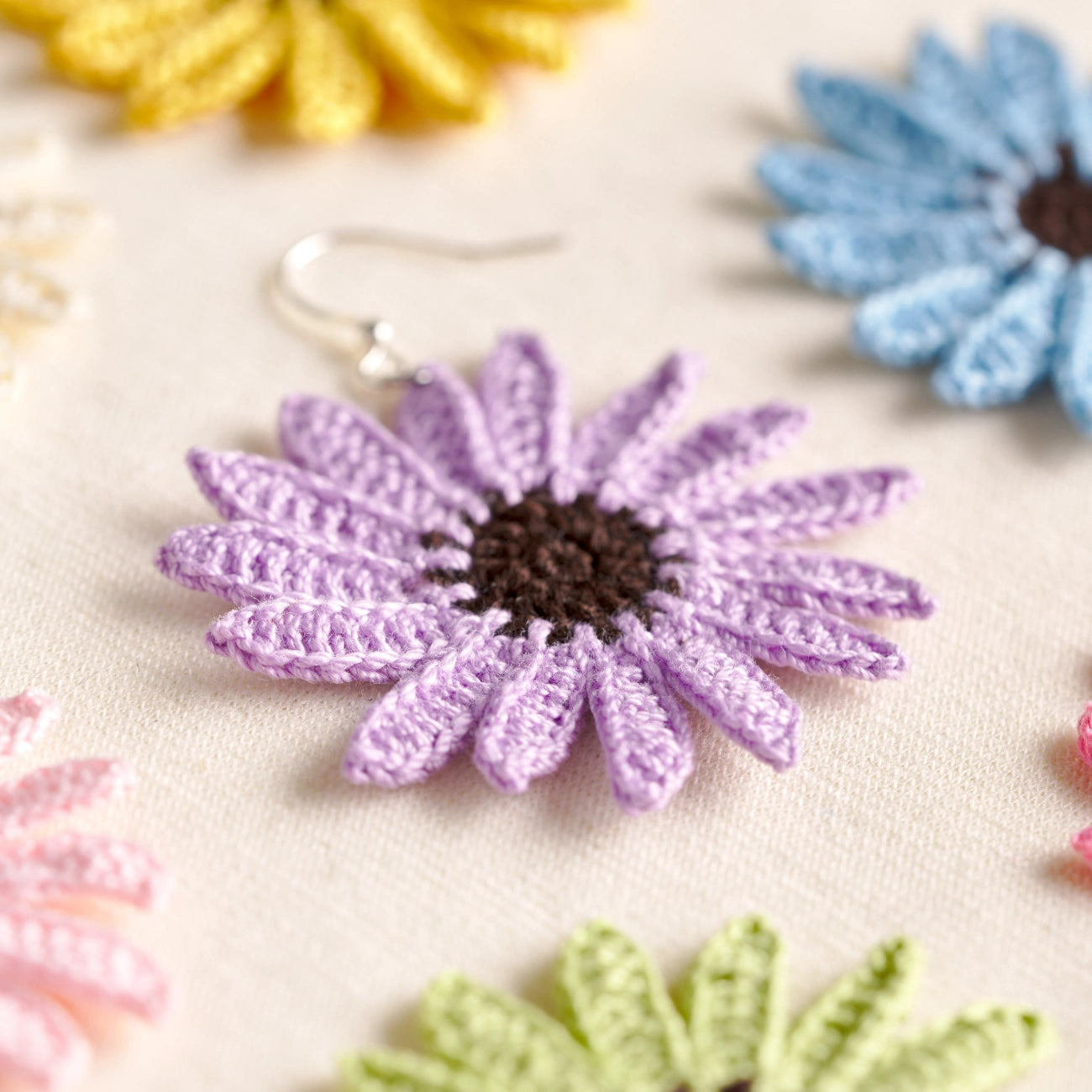 A close-up of a handmade crochet daisy earring in lavender purple with a dark brown center and silver fishhook ear wire, displayed on a light beige fabric background with other colorful crochet flowers blurred in the background.
