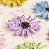 A close-up of a handmade crochet daisy earring in lavender purple with a dark brown center and silver fishhook ear wire, displayed on a light beige fabric background with other colorful crochet flowers blurred in the background.