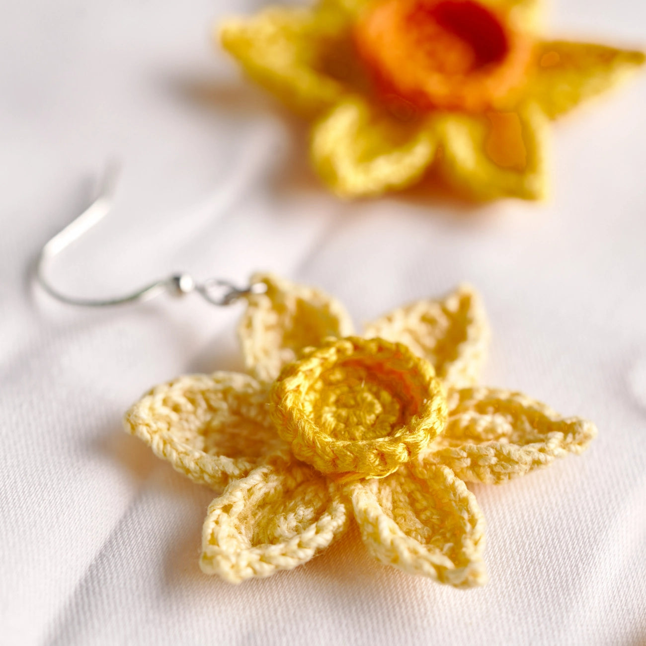 Yellow crocheted flower dangle earrings with silver fishhook wires, displayed on a white textured fabric background.
