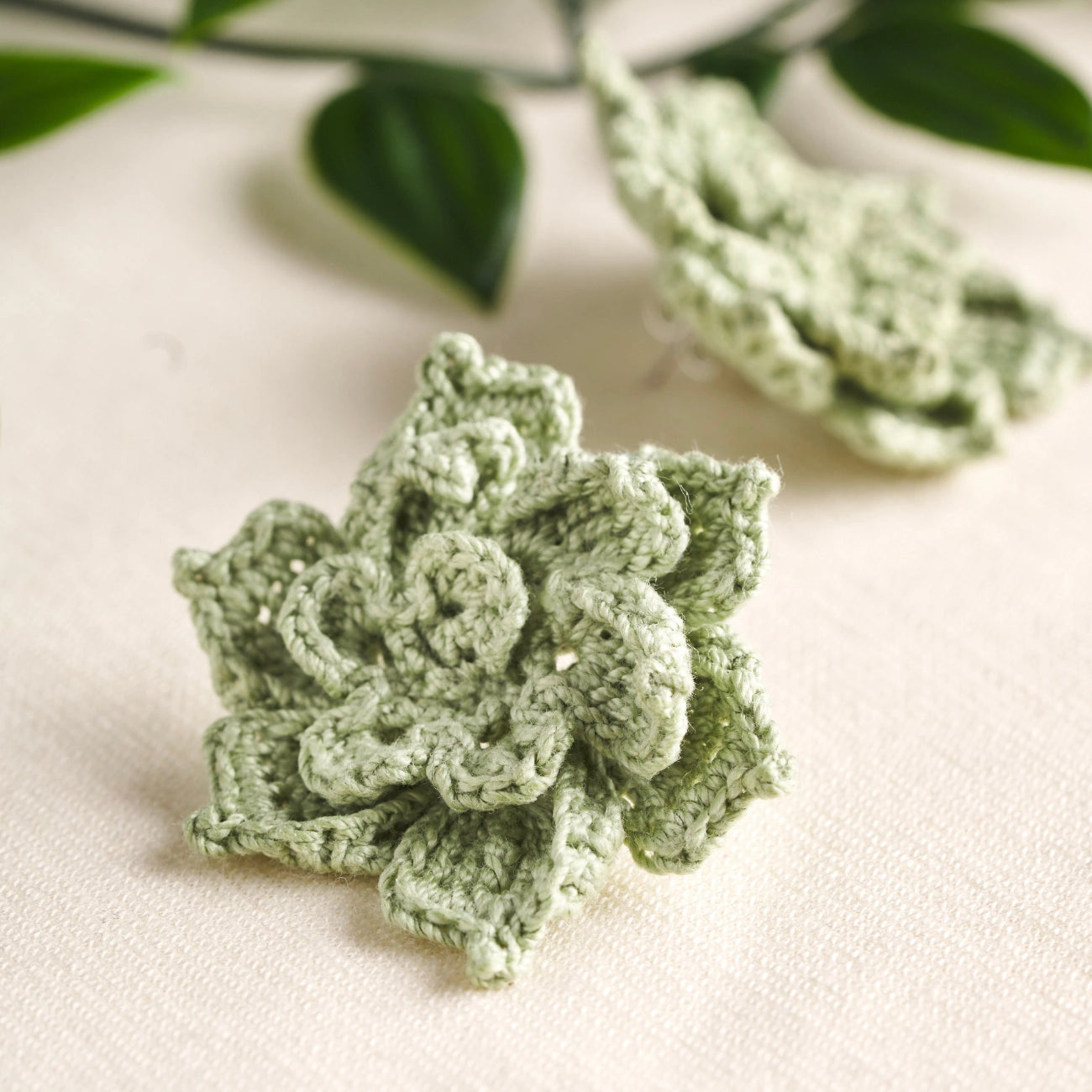A pair of light sage green crocheted flower-shaped accessories, likely hair clips or hair pins, displayed on a textured cream fabric with blurred green leaves in the background.