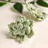 A pair of light sage green crocheted flower-shaped accessories, likely hair clips or hair pins, displayed on a textured cream fabric with blurred green leaves in the background.