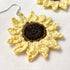 A close-up of a crocheted sunflower earring with pale yellow petals, a dark brown center, and a silver fishhook ear wire, shown on a white textured fabric background.