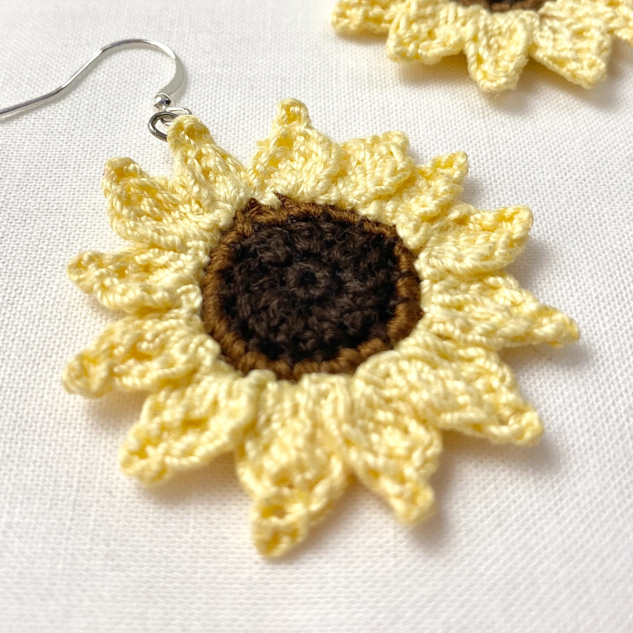 A close-up of a crocheted sunflower earring with pale yellow petals, a dark brown center, and a silver fishhook ear wire, shown on a white textured fabric background.