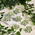 Five light sage green crochet succulent flower charms arranged on a white hexagonal dish and scattered on a beige fabric background, surrounded by green artificial vines.