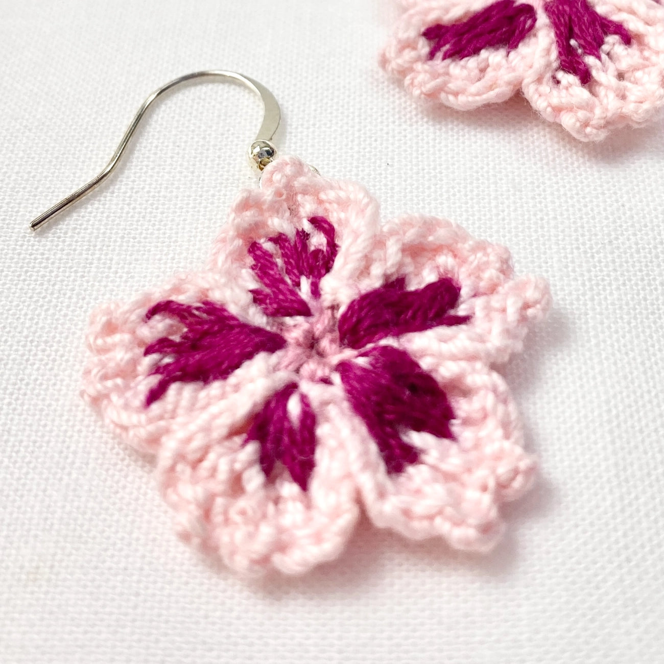 A close-up of a handmade crocheted flower earring in light pink with dark magenta embroidered petals and a silver fishhook wire, displayed on a white textured fabric background.