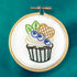 A wooden embroidery hoop holding white fabric with a colorful cupcake design. The cupcake has blueberries, green leaves, a waffle-patterned topping, and a striped liner, photographed against a teal background.