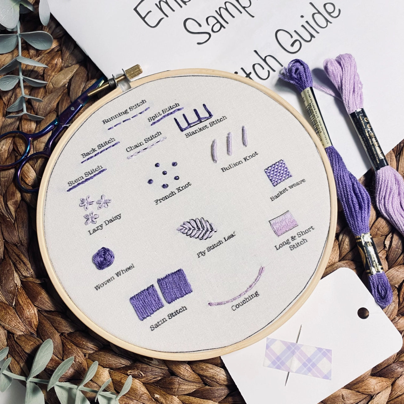 A round embroidery hoop displaying a stitch guide with labeled embroidery stitches in purple thread, surrounded by matching purple embroidery floss, a woven mat, and part of an 'Embroidery Stitch Guide' sheet.