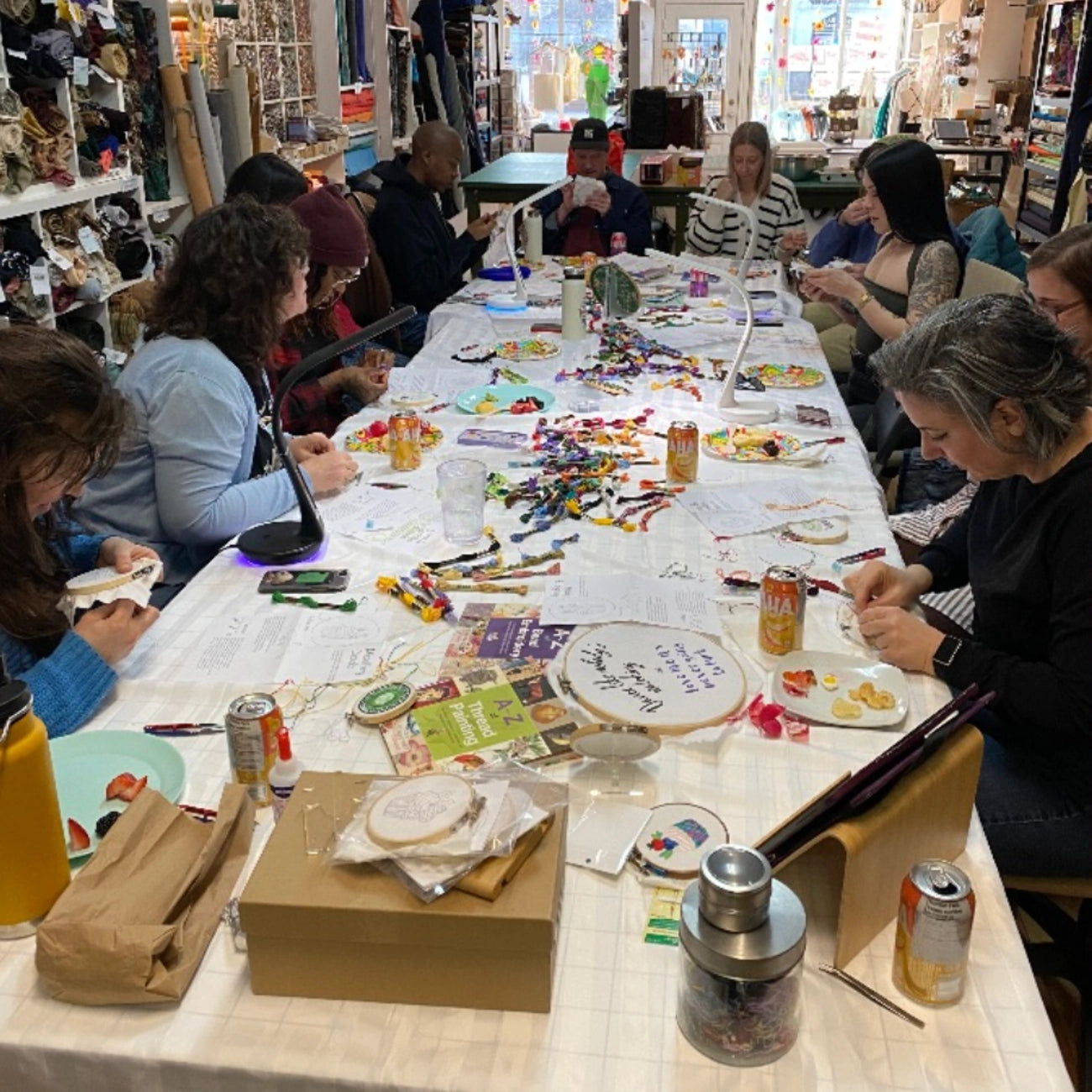 A group of people seated around a long table in a craft shop at an embroidery workshop. The table is covered with fabric, thread, needles, scissors, and small craft supplies.