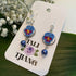 A pair of dangling earrings displayed on a white branded card. Each earring has a silver fishhook, a round blue and purple flower charm with a yellow center, a small silver bead, and a dangling blue bead.