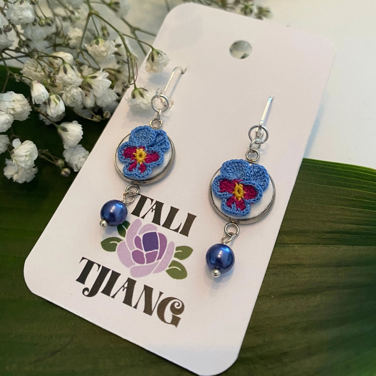 A pair of dangling earrings displayed on a white branded card. Each earring has a silver fishhook, a round blue and purple flower charm with a yellow center, a small silver bead, and a dangling blue bead.