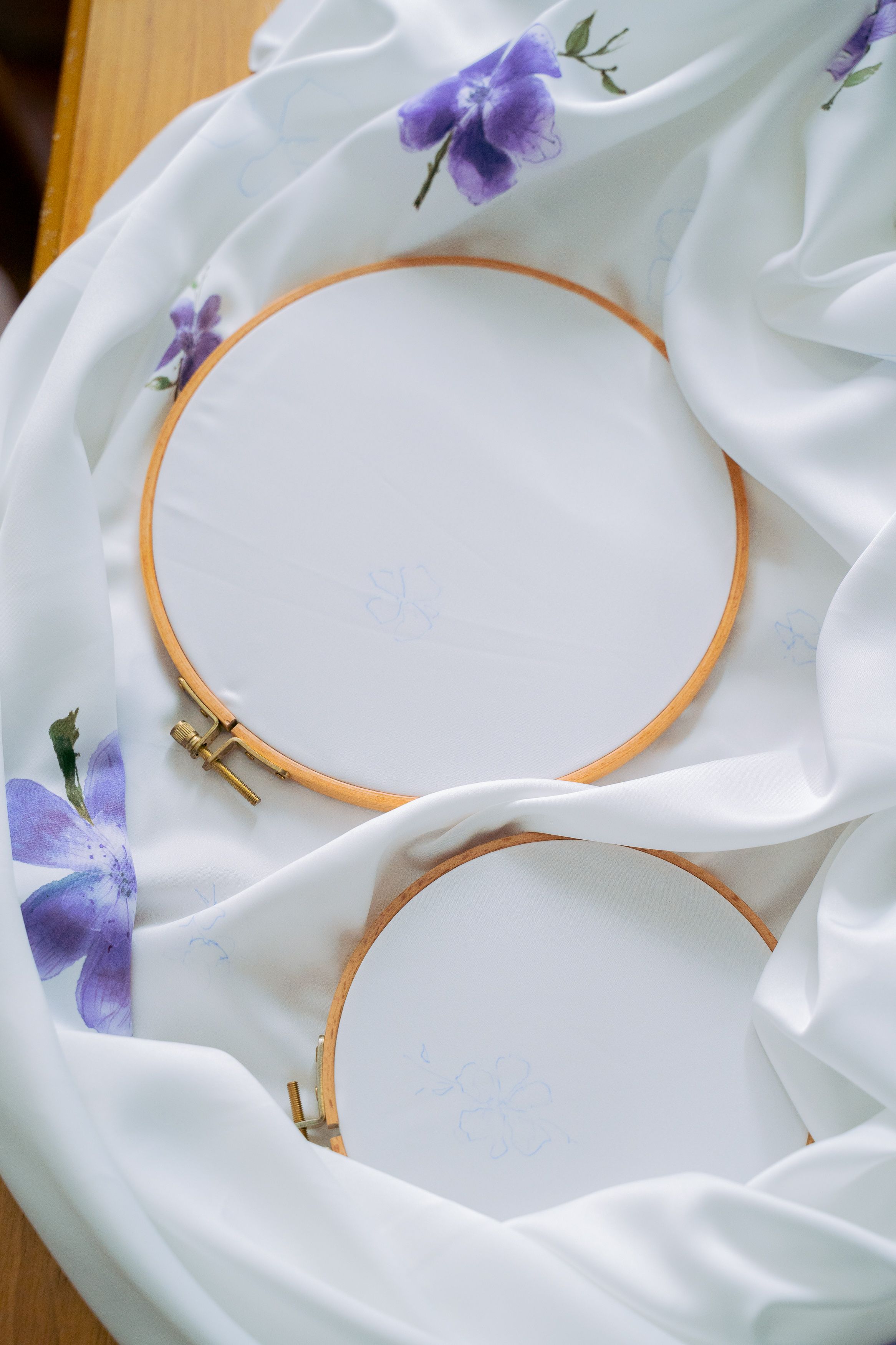 Two embroidery hoops with white fabric inserts, arranged on draped white satin with purple flowers, on a wooden surface.