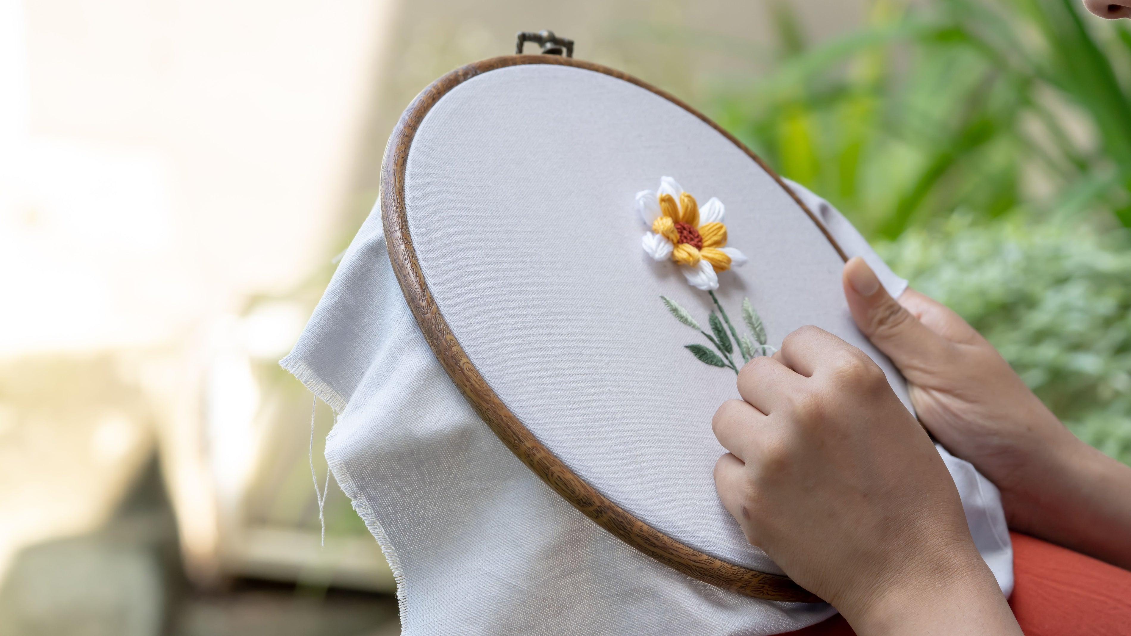 A close-up of hands holding a wooden embroidery hoop with white fabric inside, stitching a small white and yellow flower with green stems. The background is softly blurred with warm light and greenery.