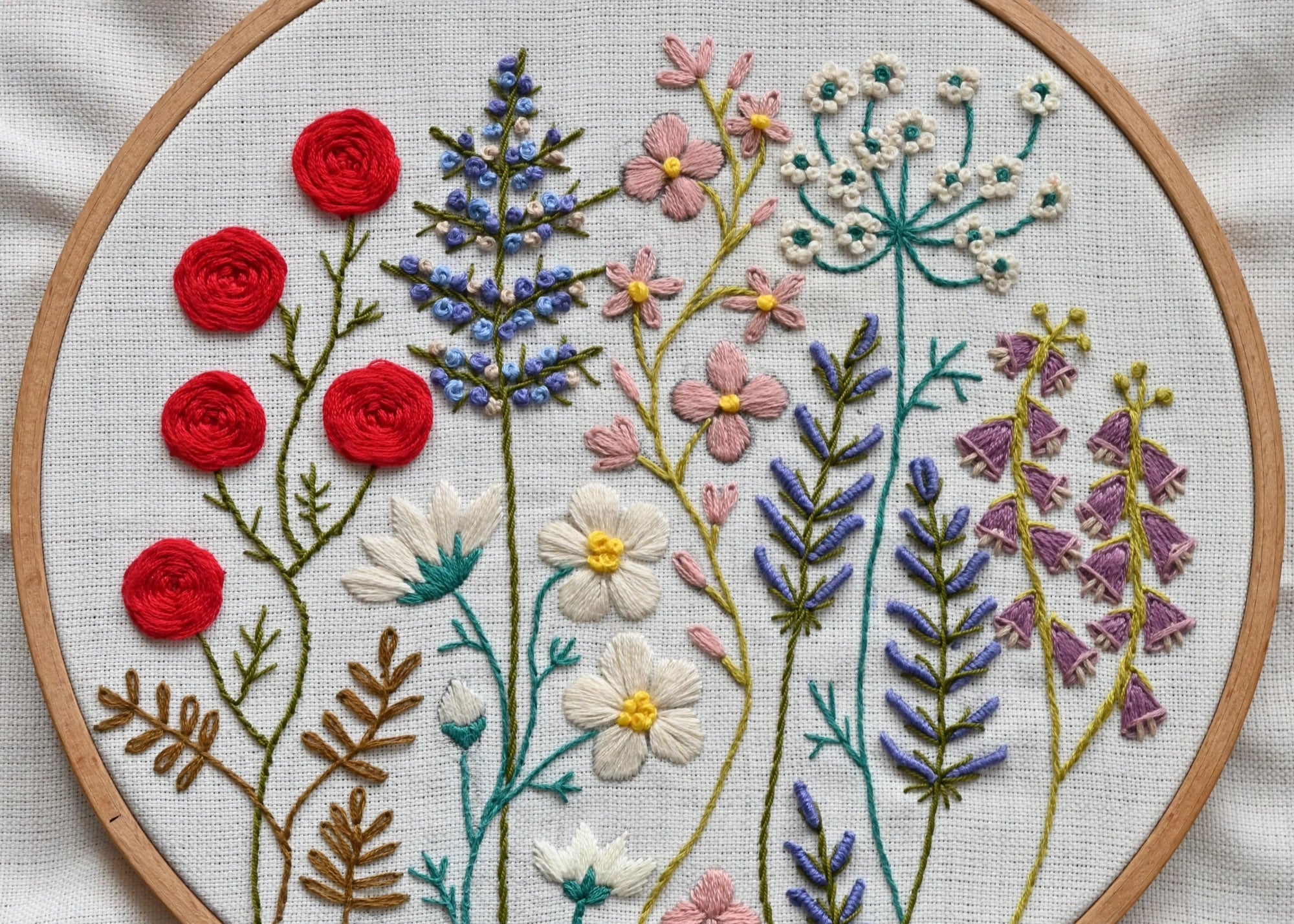 A circular embroidery hoop displays a colorful floral tapestry embroidery on white linen fabric, featuring red roses, blue lavender, pink flowers, and white daisies.