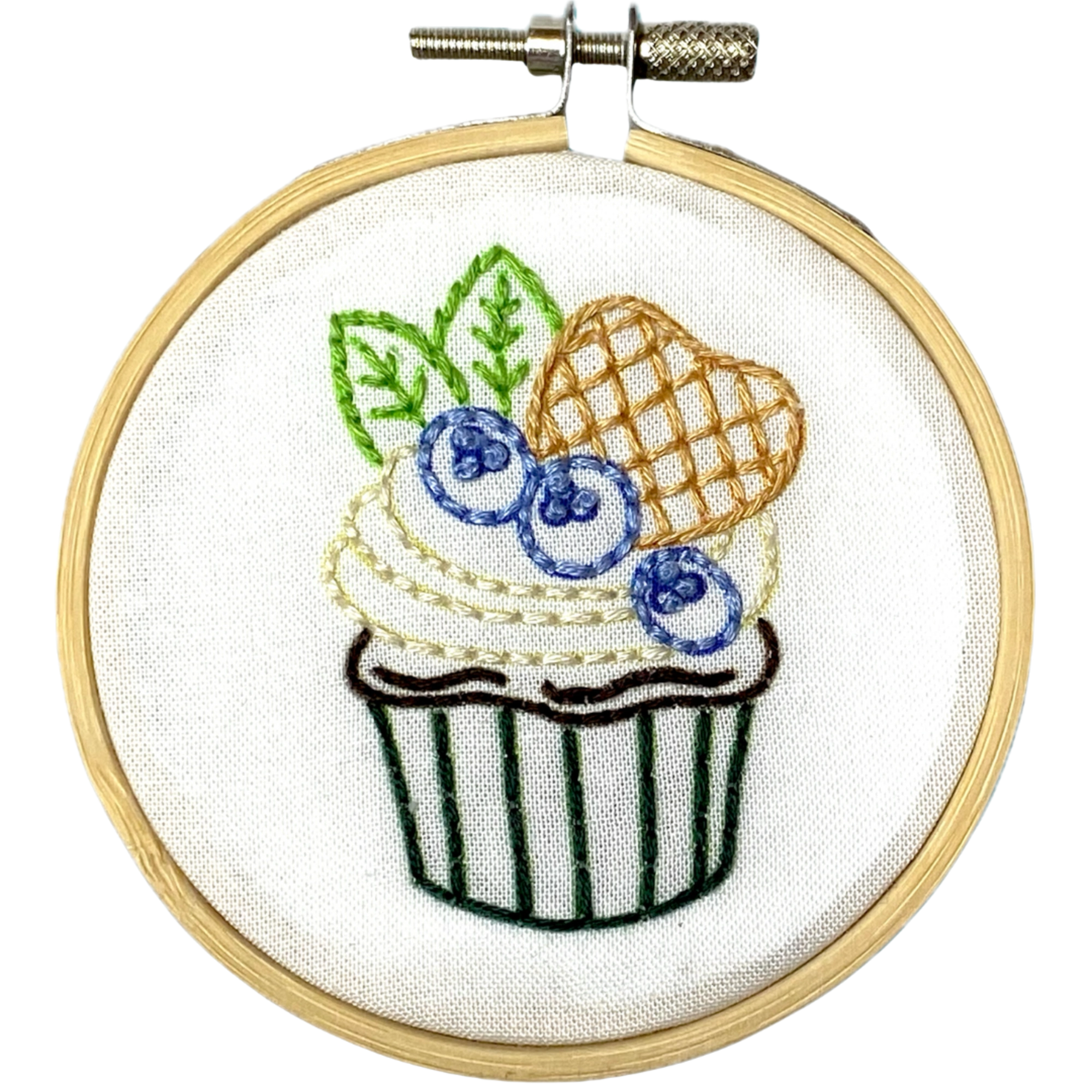 A circular embroidery hoop holding white fabric with a stitched cupcake design featuring blueberries, green leaves, a striped cupcake liner, and a waffle-patterned topping.