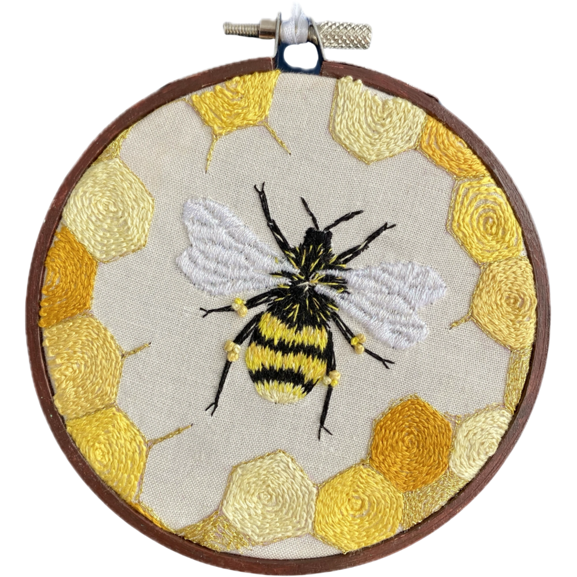 A circular embroidery hoop containing a detailed bumblebee stitched in black, yellow, and white thread, surrounded by yellow and gold hexagonal honeycomb patterns on a light beige fabric background.
