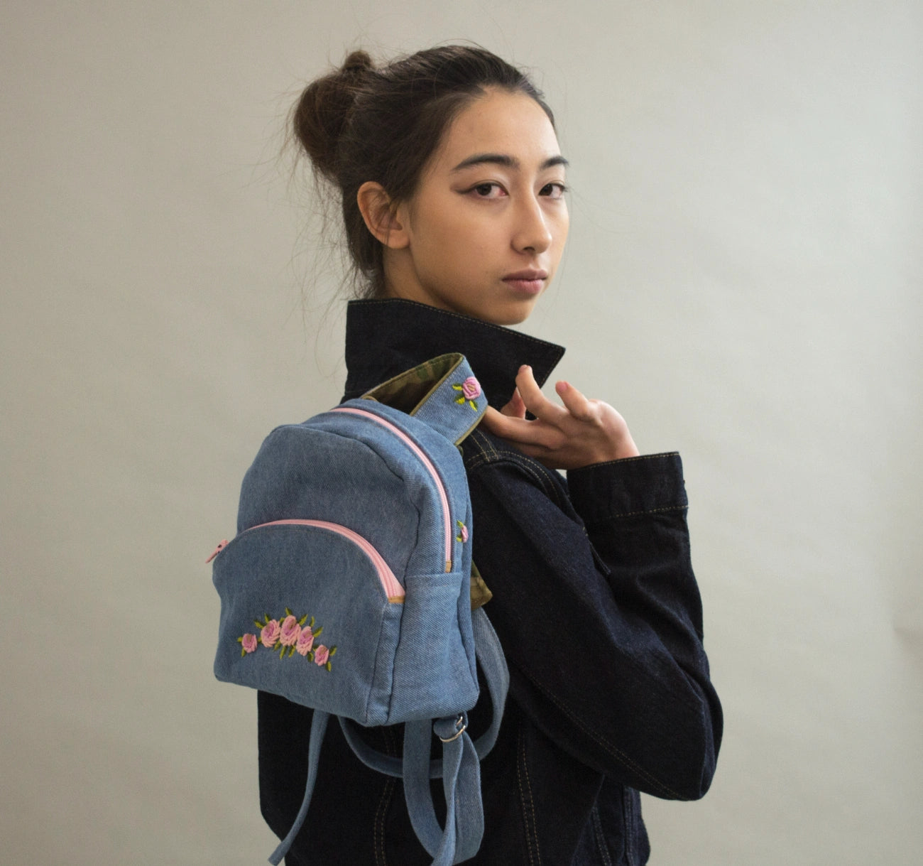 A model wearing a dark denim jacket and black turtleneck, carrying a small blue denim backpack with pink zipper accents and floral embroidery on the front pocket.