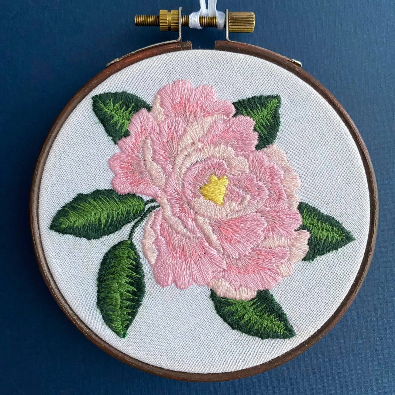 A close-up of a floral embroidery piece in a wooden hoop, featuring a large pink flower with green leaves and a yellow center on white fabric, set against a dark blue background.