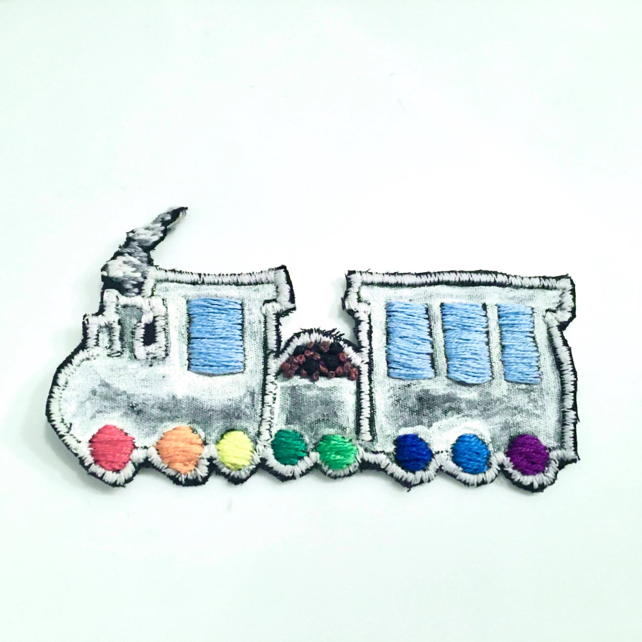 An embroidered patch shaped like a cartoon train, featuring a gray body with blue windows and a row of colorful wheels in pink, orange, yellow, green, blue, and purple on a white background.