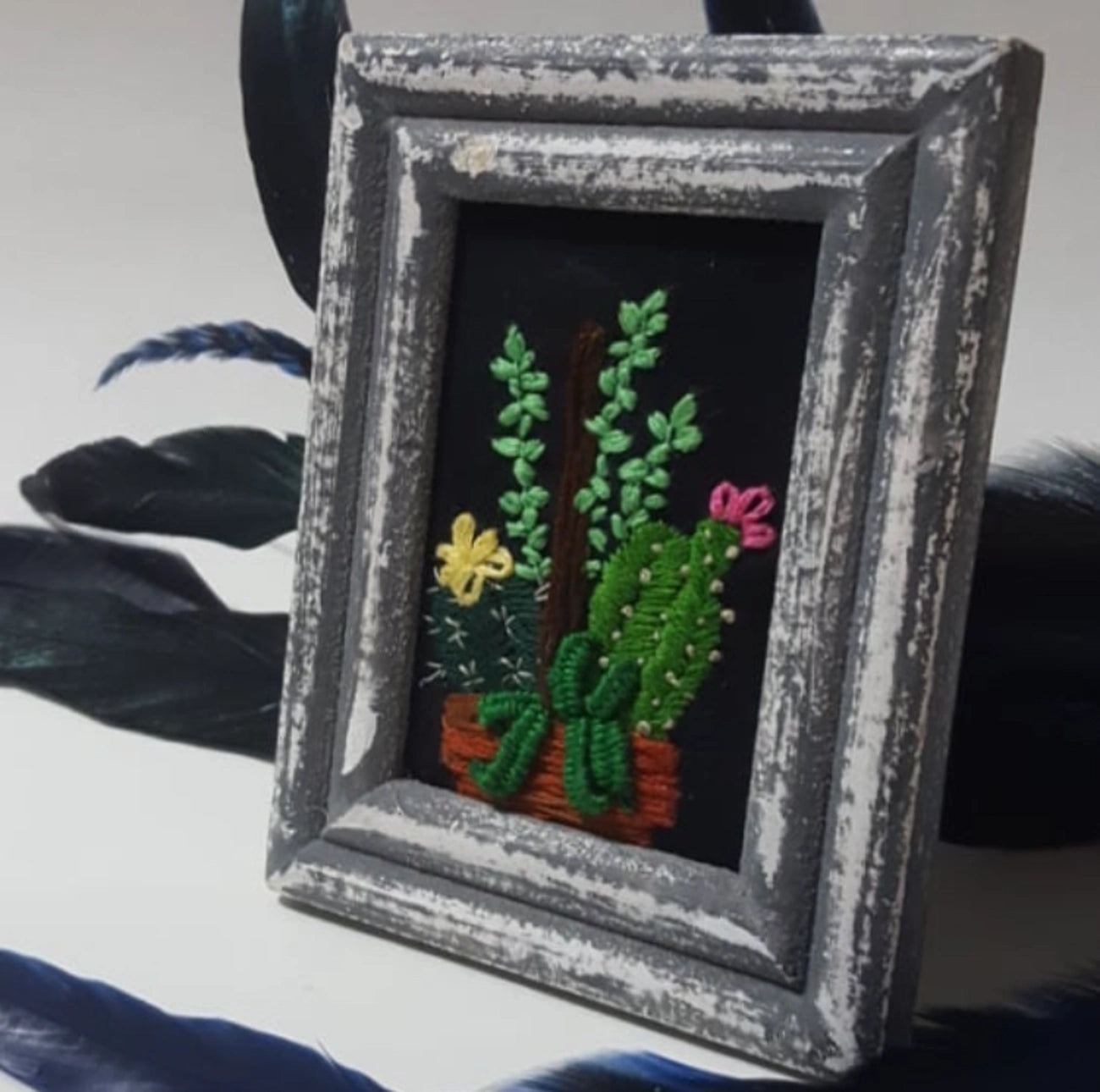 A small framed embroidered cactus and succulent artwork in a distressed gray frame, set against a white background with dark feathers.