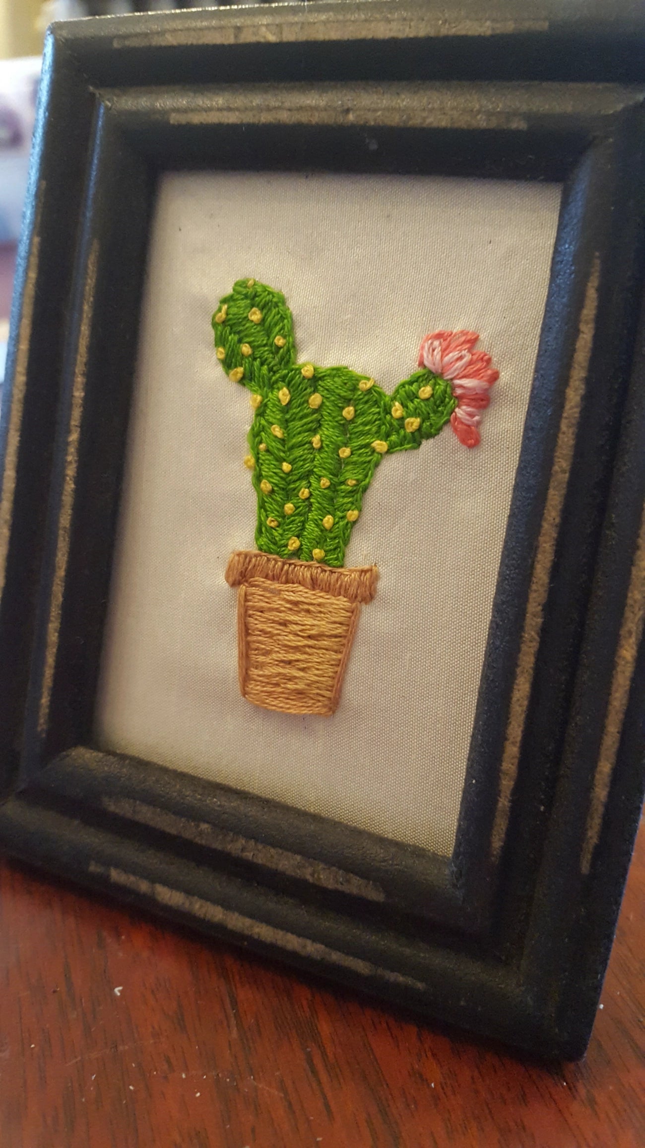A small framed embroidered cactus artwork on a light fabric background, placed on a wooden surface. The cactus is green with yellow stitched dots, a tan pot, and a pink flower, all enclosed in a dark wooden frame.