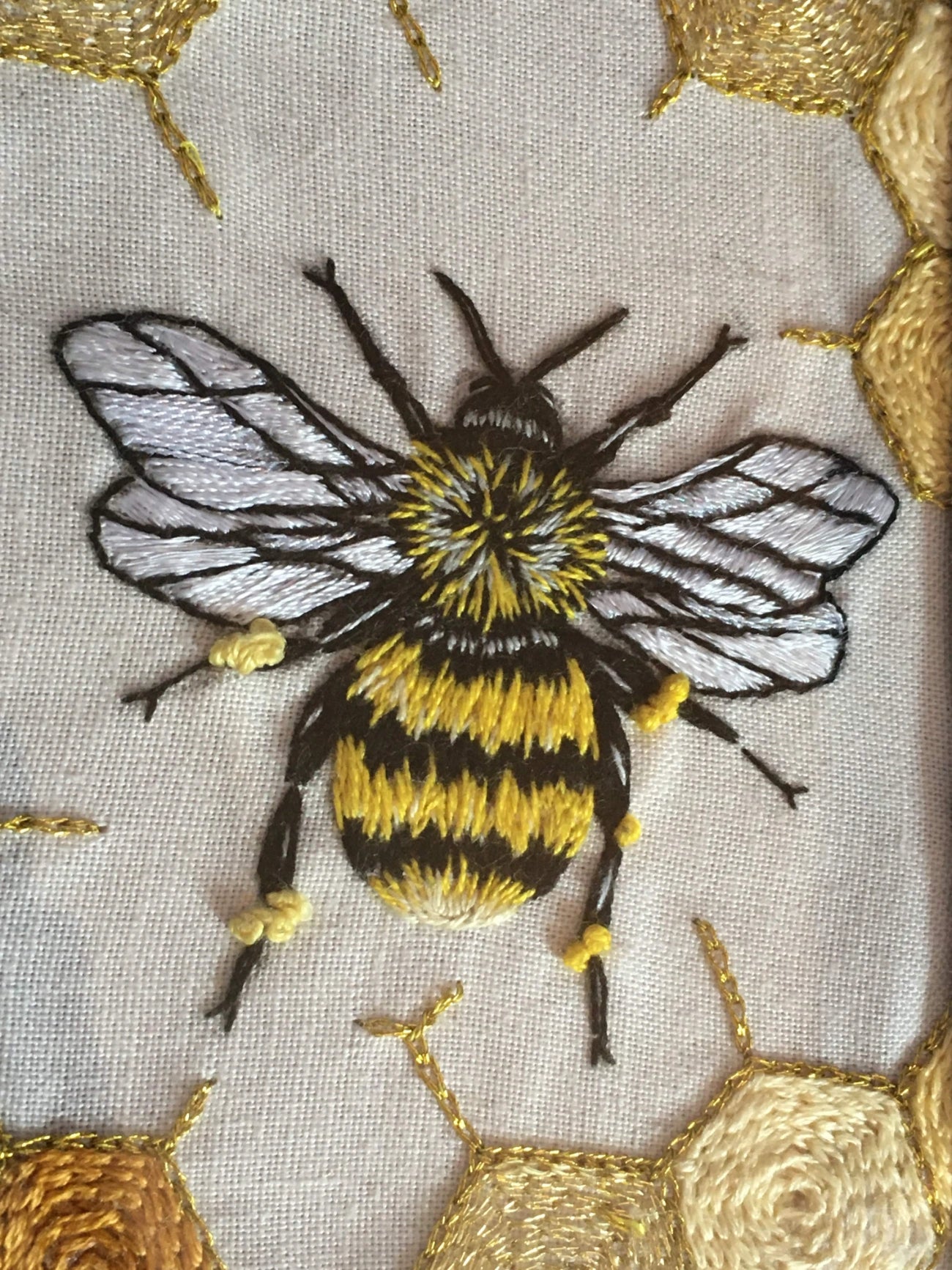 Close-up of an embroidered bumblebee motif on a light beige fabric background with gold hexagonal stitching.