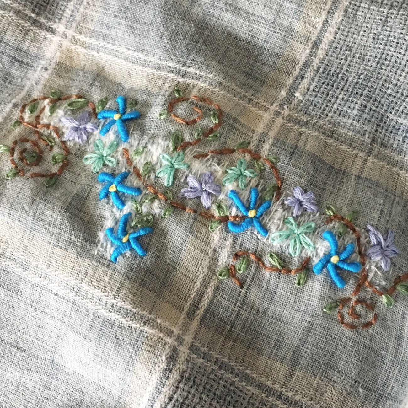 A close-up of a textured beige and gray plaid fabric featuring delicate embroidered floral details in blue, green, purple, and brown.