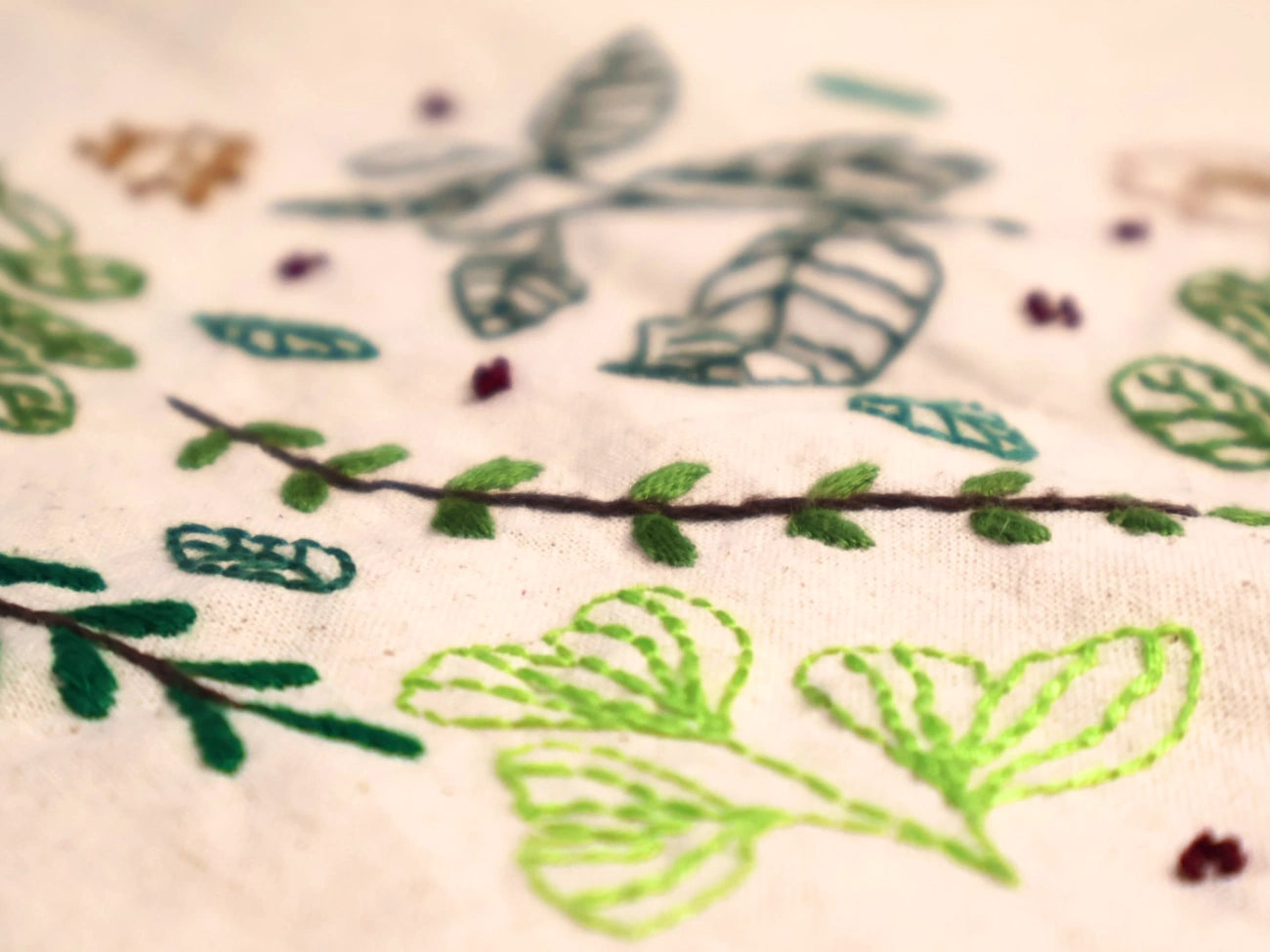 Close-up of embroidered botanical leaf patterns in green, teal, and brown on a light cream fabric background.