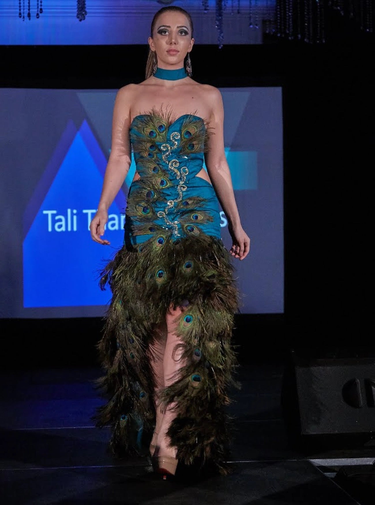 A model walks a fashion runway wearing a strapless teal gown with peacock feather detailing and a matching feathered skirt.
