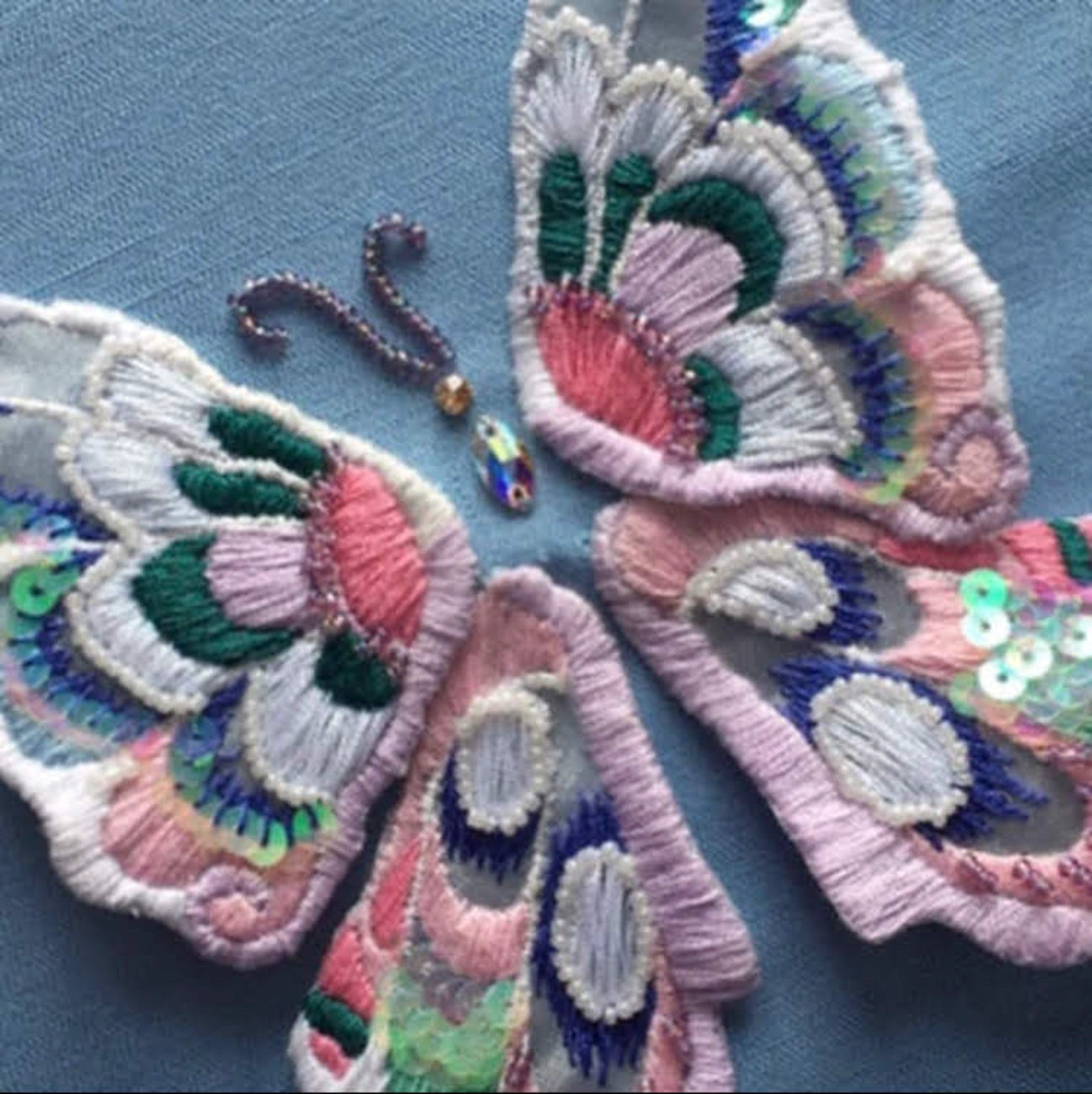 A set of four butterfly wing-shaped embroidered patches in pastel pink, green, blue, and white, arranged on a textured blue background.