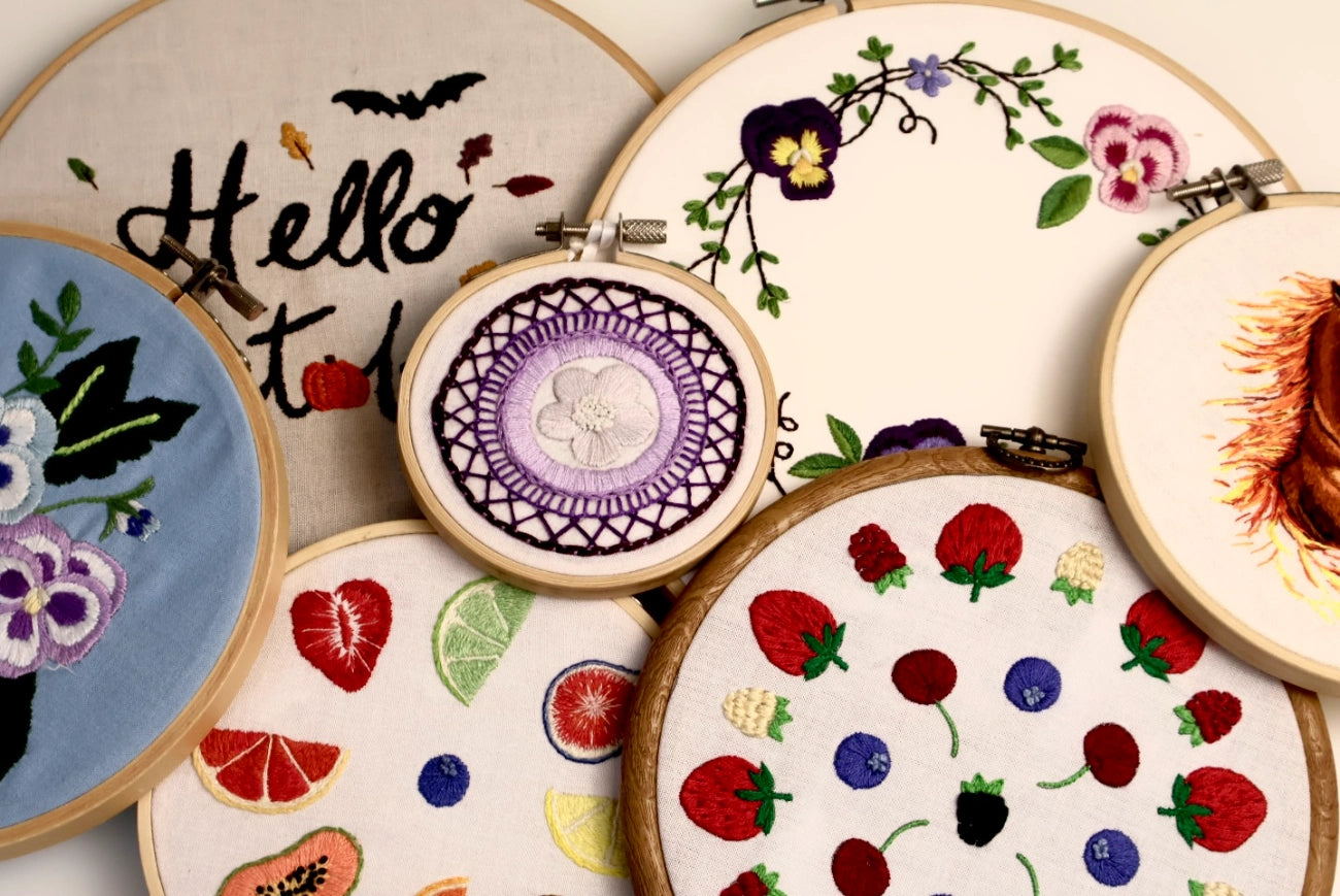 A collection of wooden embroidery hoops displaying various colorful floral and fruit embroidery designs on cream fabric.