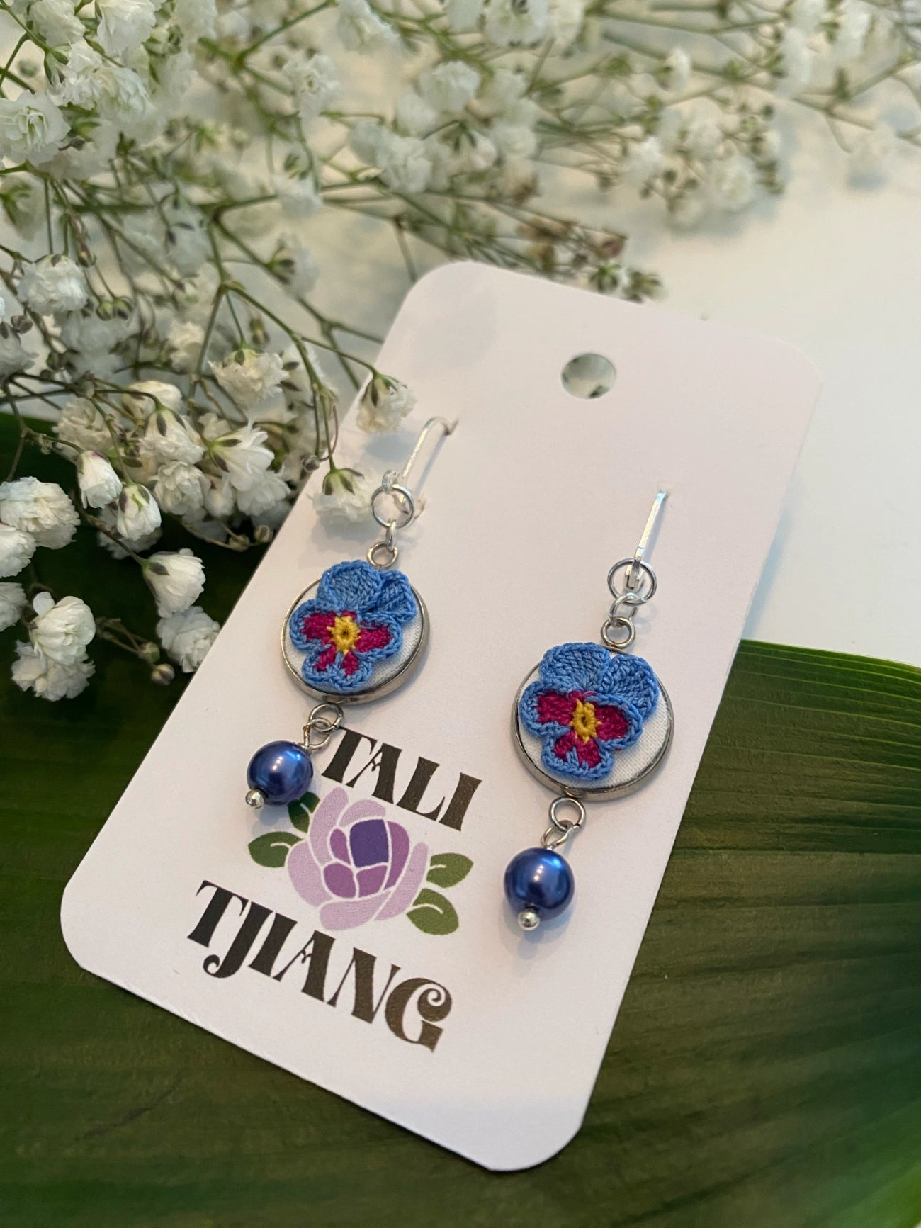 A pair of handmade dangle earrings displayed on a white brand card. The earrings feature a round blue and purple flower charm with a yellow center, a silver-tone metal ring, and a dangling blue bead.