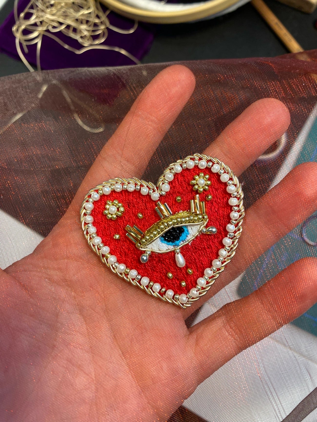 A heart-shaped red embroidered patch with a gold crown, white pearl border, and a central blue eye, held on a person's hand.